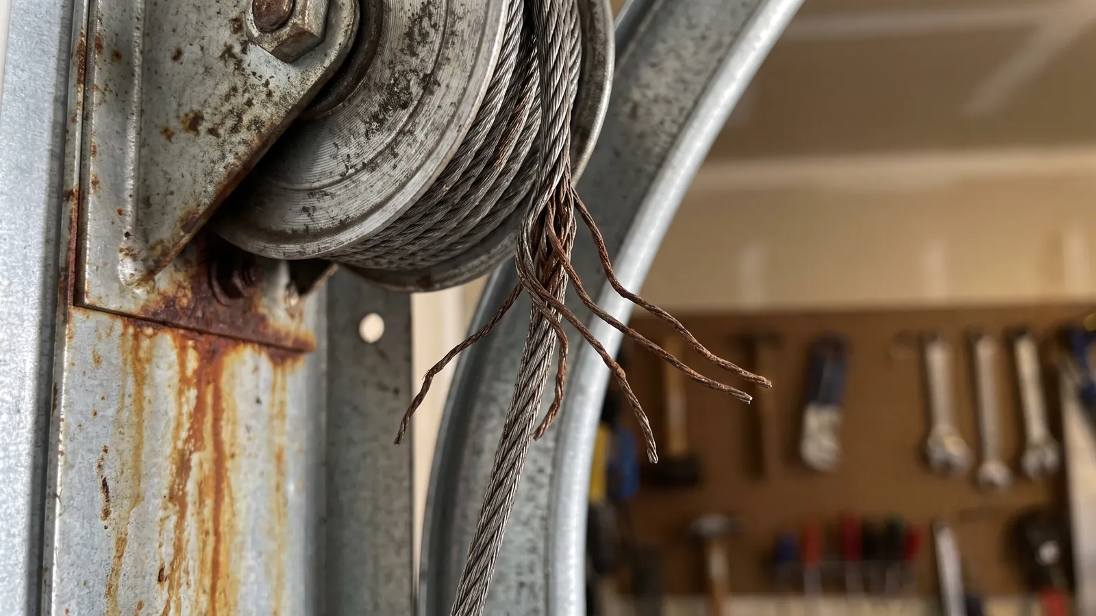 Close-up of a frayed steel lift cable unwound from its grooved cable drum at the top corner of a garage door track